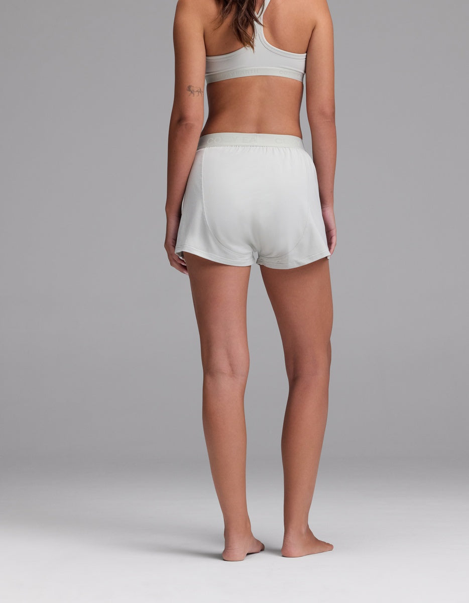A woman stands barefoot on a plain surface, facing away. She wears the Cozy Earth Women’s First Layer Pajama Boxer Short and a light gray sports bra. The background is neutral gray.