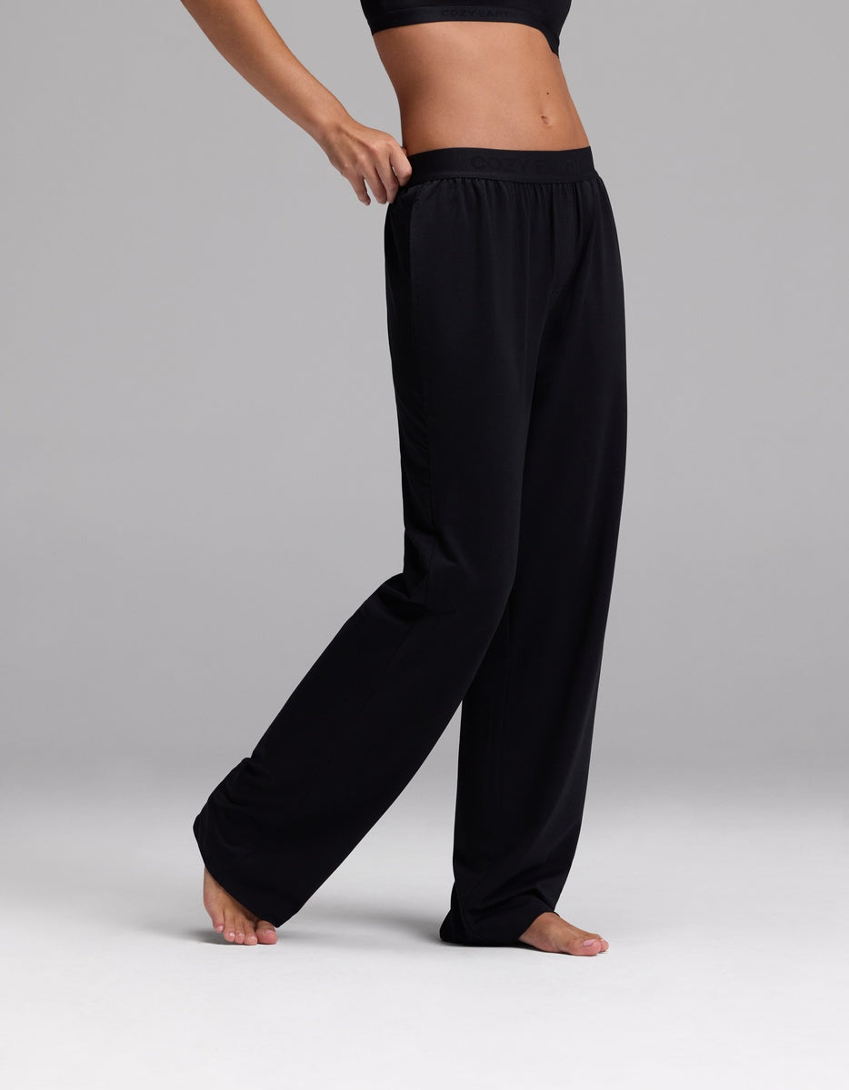 A person wearing Cozy Earth Women’s First Layer Pajama Pant in Jet Black stands barefoot on a light gray surface, posing with one hand on their hip and one leg slightly bent.|Color:Jet Black