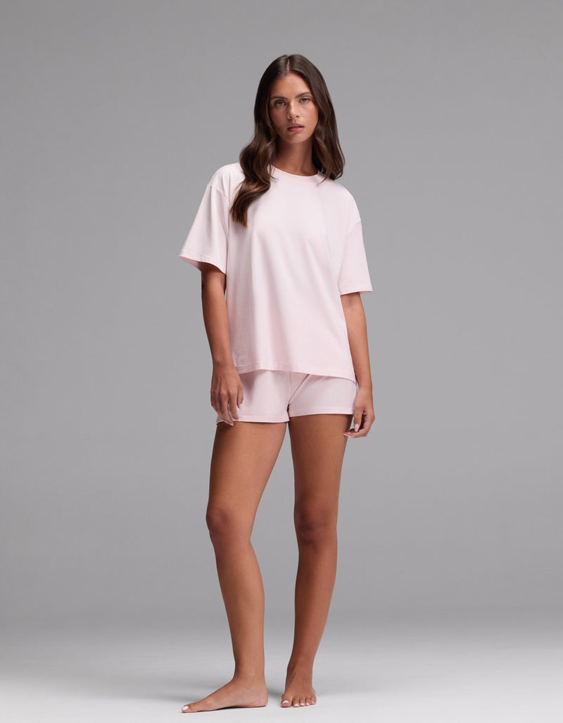 A woman stands barefoot against a plain background, wearing the Cozy Earth Women’s First Layer Pajama Short Sleeve Tee & Boxer Short Set in Ballet. She has long brown hair and a neutral expression.