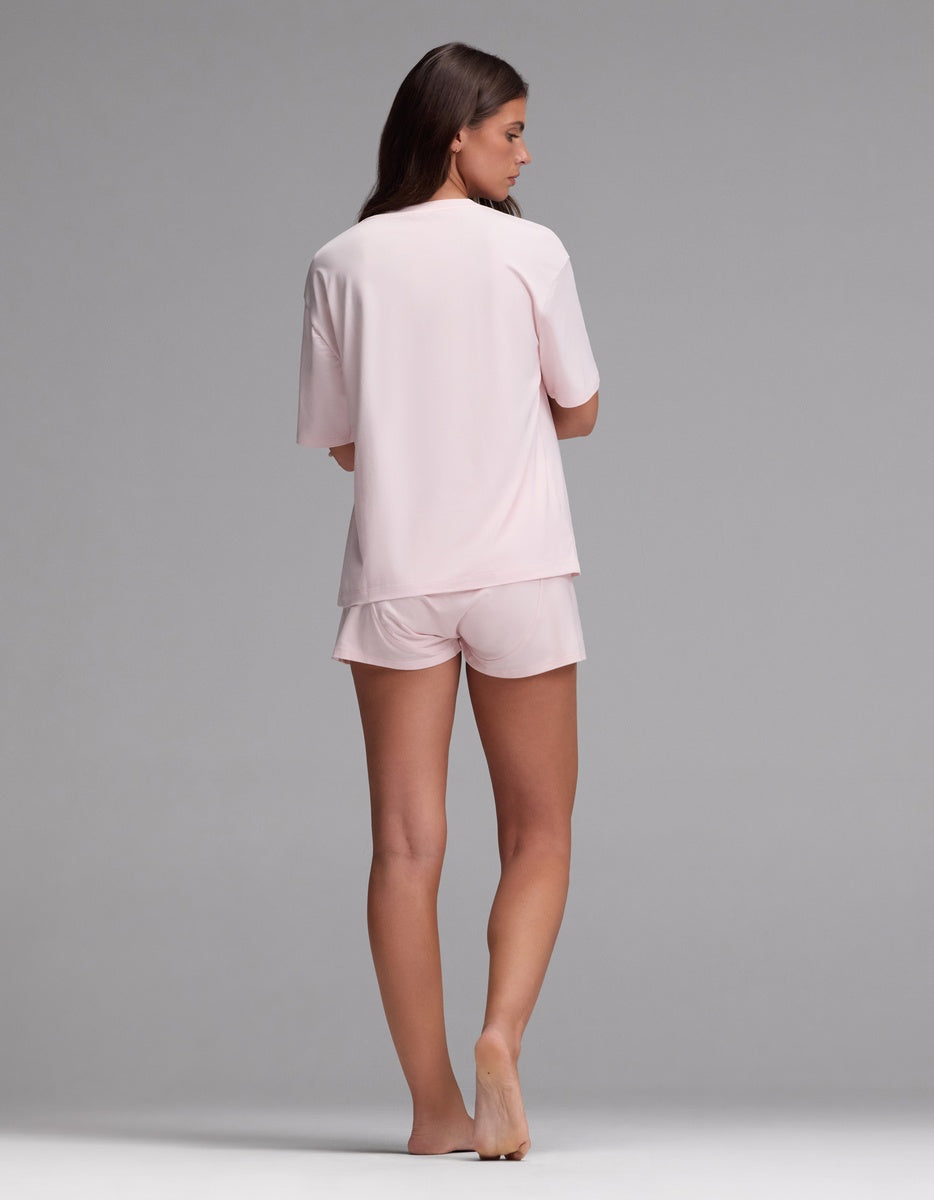 A woman stands barefoot facing away, wearing the Cozy Earth Women’s First Layer Pajama Short Sleeve Tee & Boxer Short Set in light pink, against a plain gray background.