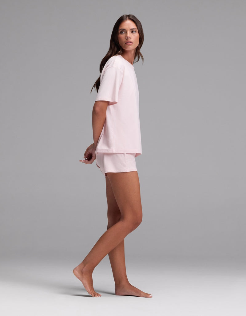 A woman stands barefoot in profile, wearing the Cozy Earth Women’s First Layer Pajama Short Sleeve Tee & Boxer Short Set in light pink against a plain gray background.