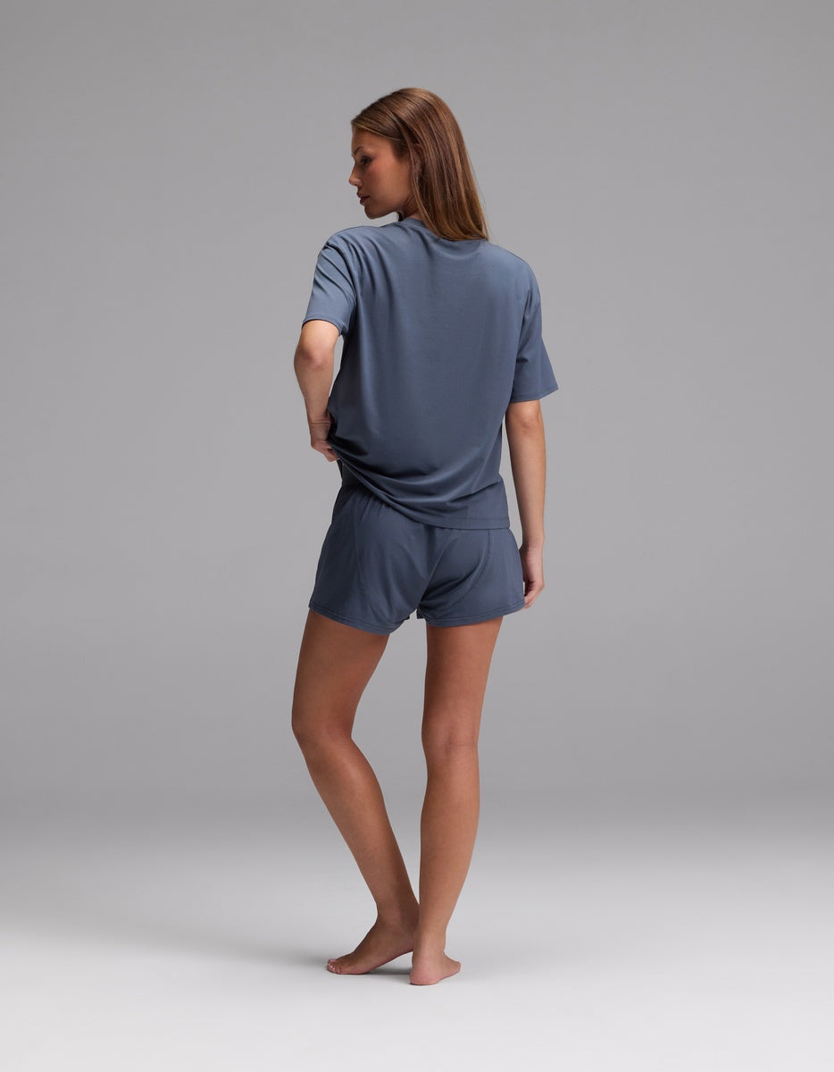 A woman, barefoot and facing away, models the Cozy Earth Women’s First Layer Pajama Short Sleeve Tee & Boxer Short Set in gray against a plain light gray background.