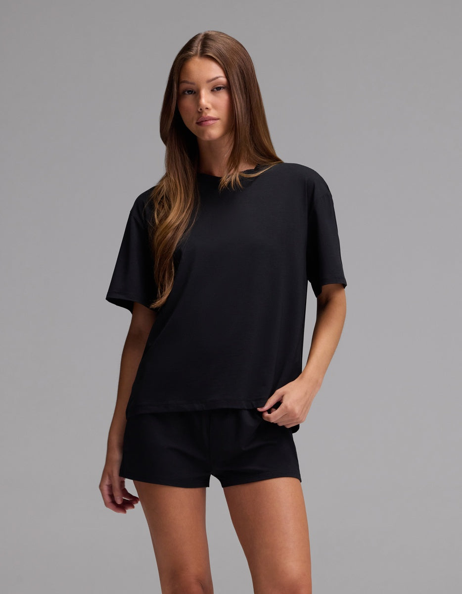 A woman stands against a plain gray background, wearing the Cozy Earth Women’s First Layer Pajama Short Sleeve Tee & Boxer Short Set in black. She has long, straight brown hair and a relaxed expression, with one hand resting in her shorts pocket.