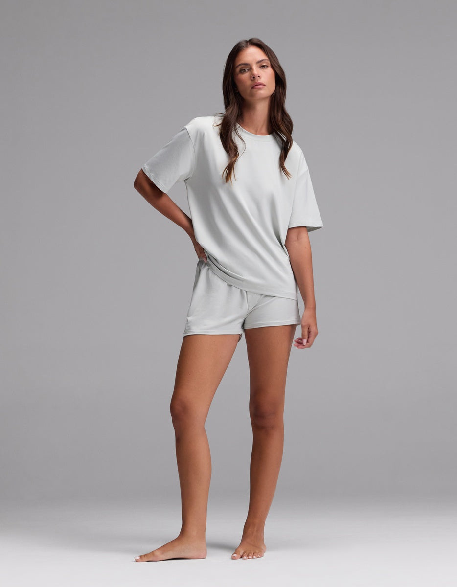 A woman stands barefoot against a plain background, wearing the Women’s First Layer Pajama Short Sleeve Tee & Boxer Short Set by Cozy Earth in Laurel, with a relaxed pose and neutral expression.