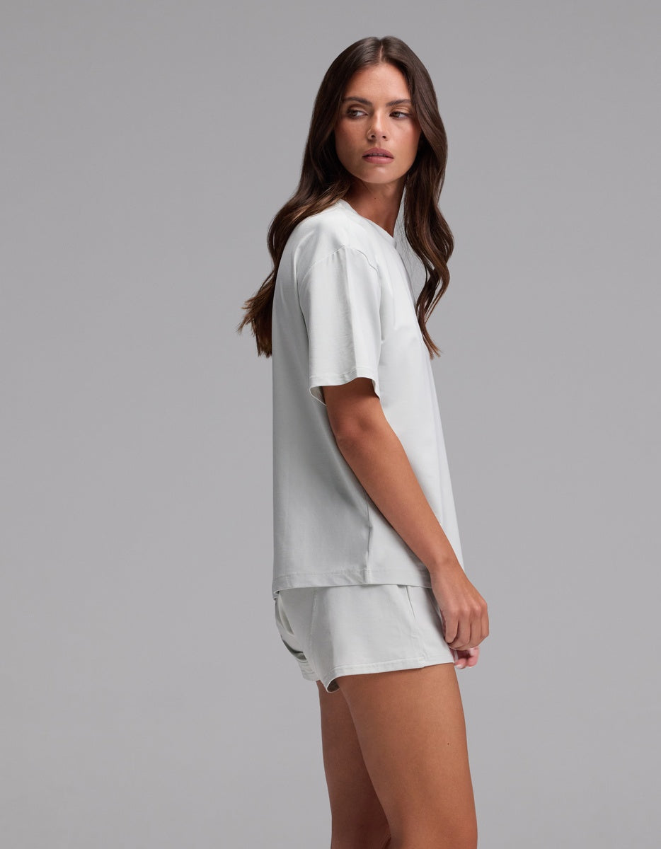 A woman with long brown hair stands sideways against a plain gray background, wearing the Cozy Earth Women’s First Layer Pajama Short Sleeve Tee & Boxer Short Set in light gray.