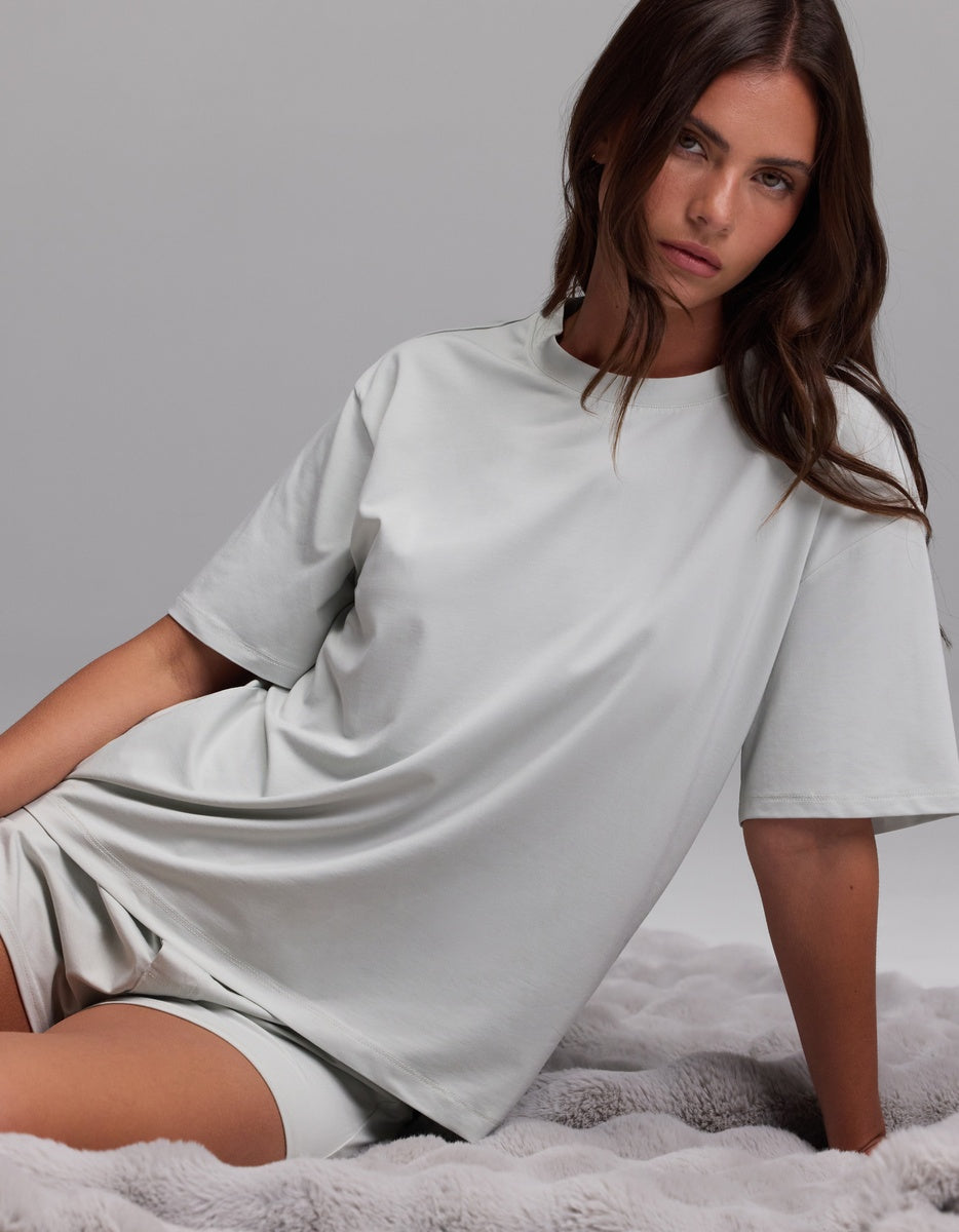 A woman with long brown hair wears the Cozy Earth Women’s First Layer Pajama Short Sleeve Tee & Boxer Short Set in light gray, sitting on a soft, textured gray blanket against a plain background.
