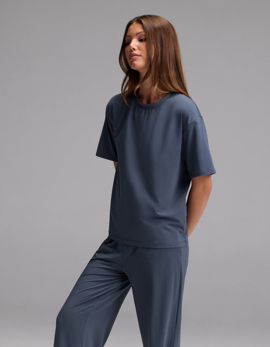 A woman with long brown hair stands against a plain gray background, wearing the Cozy Earth Women’s First Layer Pajama Short Sleeve Tee in dark blue along with matching pants. She gazes slightly to the side with a relaxed expression.|Color:Dawn