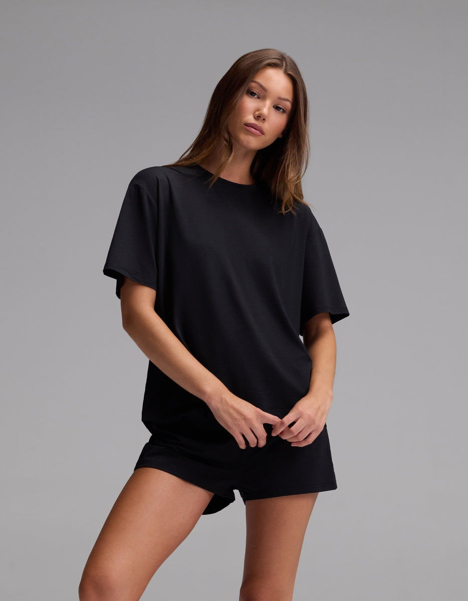A woman wearing the Cozy Earth Women’s First Layer Pajama Short Sleeve Tee and matching black shorts stands against a plain gray background, looking at the camera with a relaxed pose and neutral expression.|Color:Jet Black