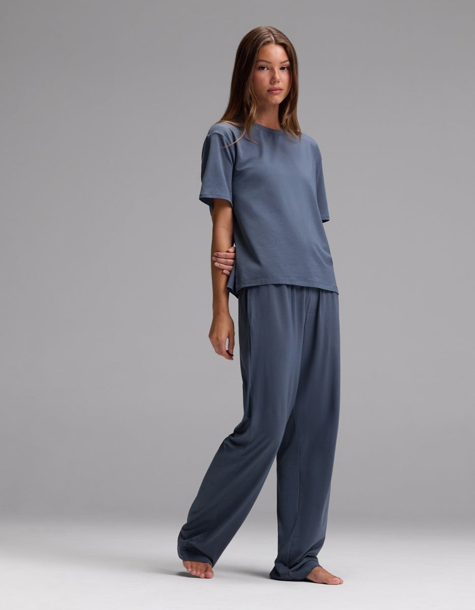 A woman stands barefoot against a plain gray backdrop wearing the Cozy Earth Women’s First Layer Pajama Short Sleeve Tee & Pajama Pant Set in Dawn, featuring a relaxed fit and muted blue hue.|Color:Dawn