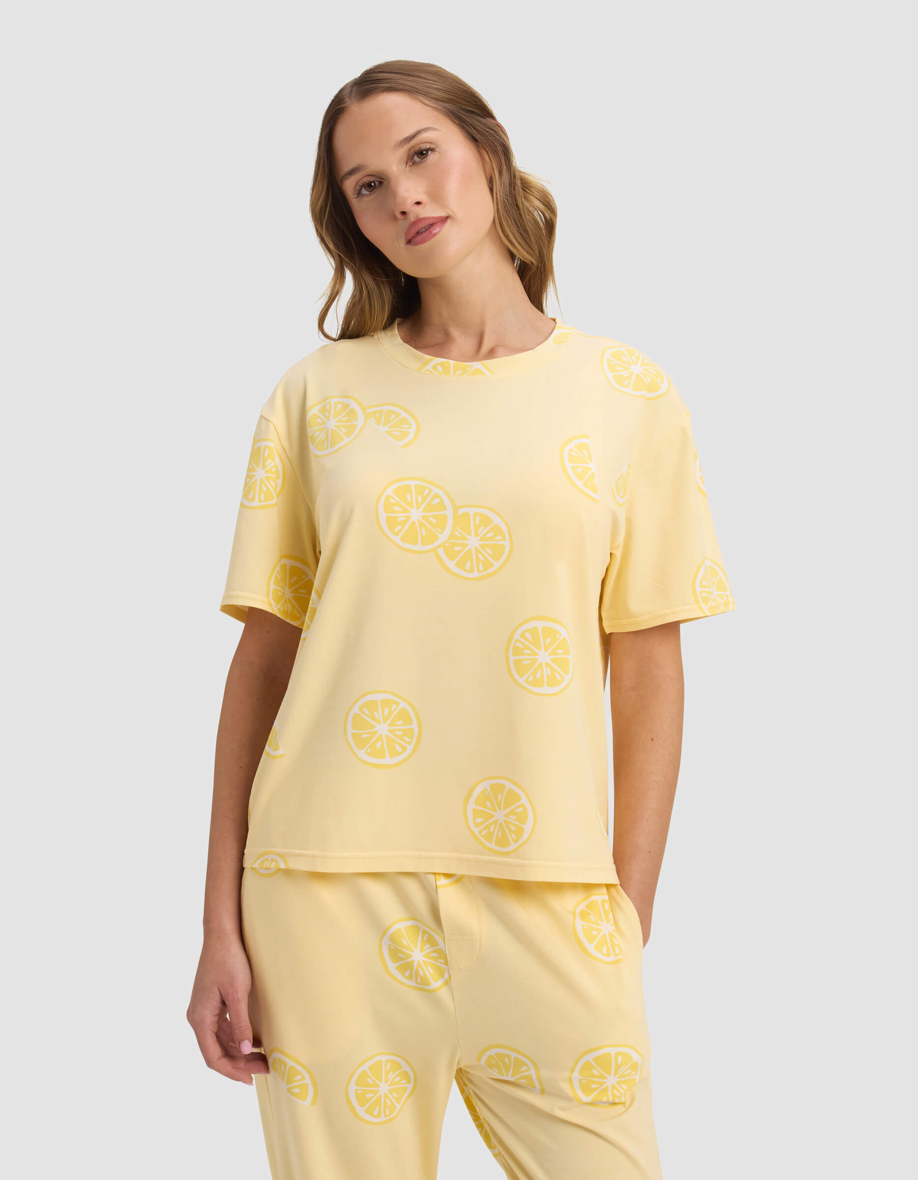 A woman wears the Cozy Earth Women's First Layer Pajama Short Sleeve Tee, paired with matching lemon slice patterned pants, standing against a plain light gray background.|Color:Lemons