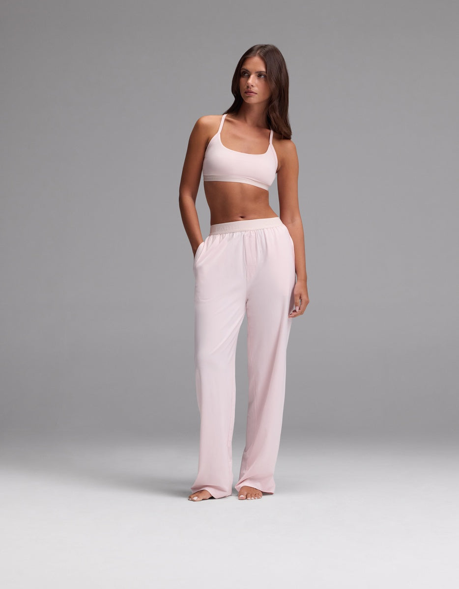 A woman stands against a plain gray background wearing the Cozy Earth Women’s First Layer Soft Bra in light pink, paired with matching loose-fitting pants, her hands in her pockets as she gazes slightly to the side.