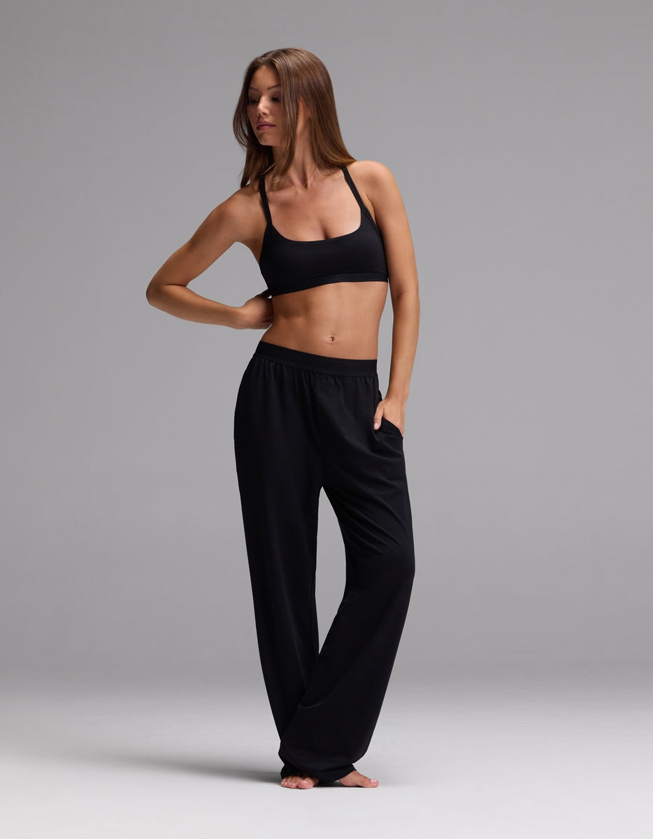 A woman models the Cozy Earth Women’s First Layer Soft Bra and loose black pants, standing barefoot on a gray background with one hand on her hip and the other resting on her thigh, gazing to the side.
