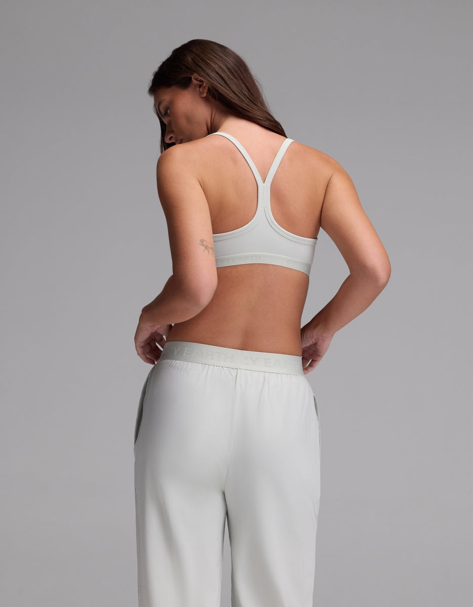 A woman with long brown hair models the Cozy Earth Women’s First Layer Soft Bra in light gray, paired with matching loose pants, standing with her back turned and looking over her shoulder against a plain gray background.