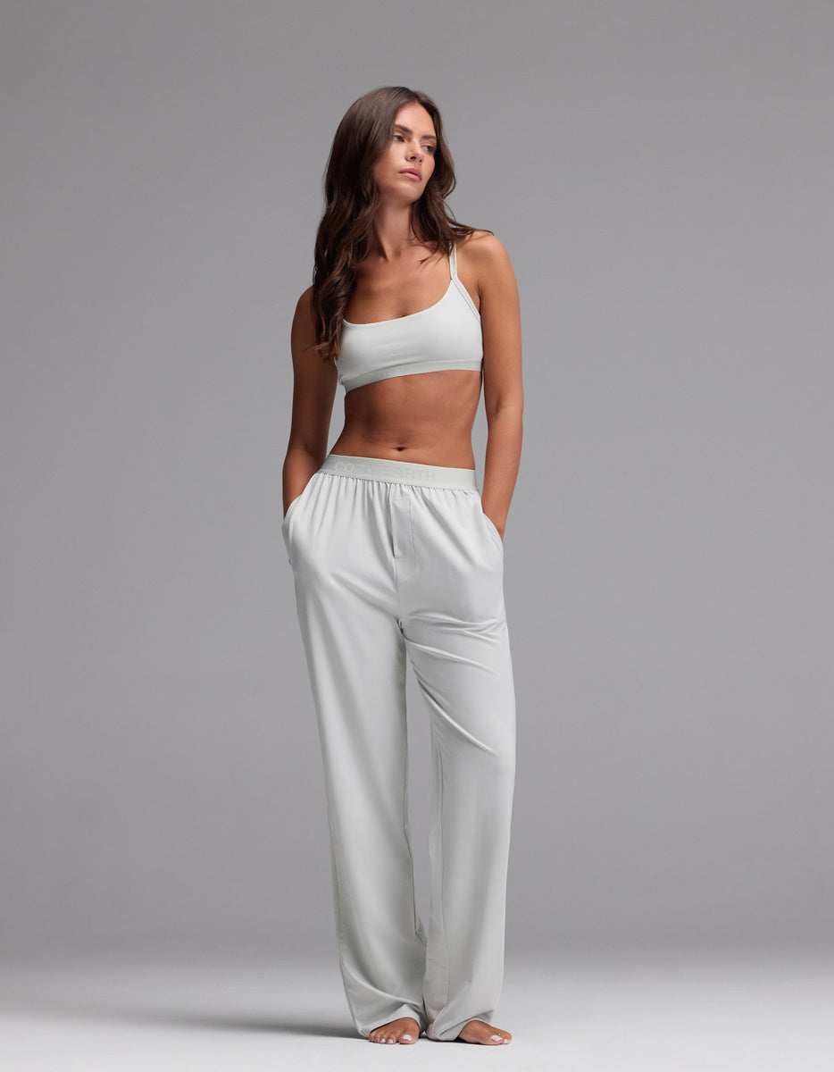 A woman stands barefoot against a gray background, wearing the Cozy Earth Women’s First Layer Soft Bra paired with matching loose pants. Her hands are in her pockets and she gazes slightly to the side with a neutral expression.