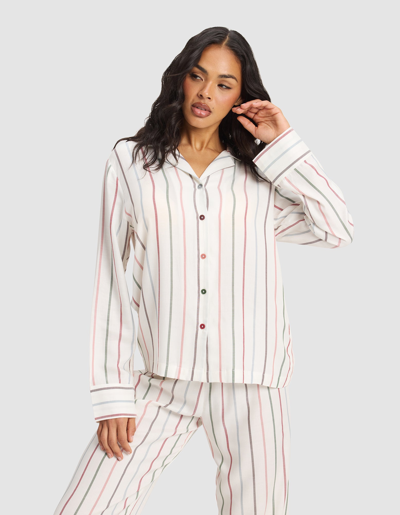 A woman with long, wavy dark hair models the Cozy Earth Women's Soft Woven Long Sleeve Pajama Set, featuring vertical red, green, and beige stripes on white, against a plain light gray background in a relaxed pose.