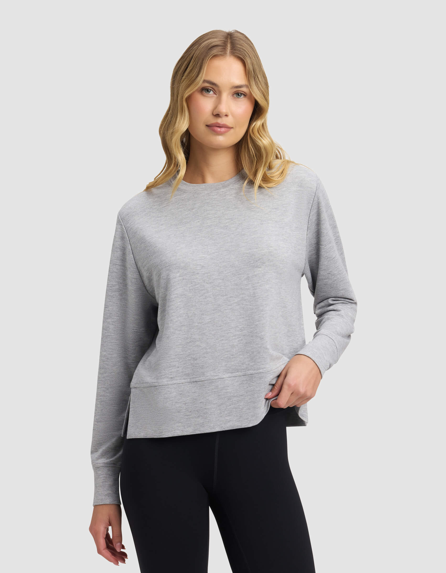 A woman with wavy blonde hair wears the Cozy Earth Women's StudioLite Crewneck in light gray with black leggings, standing against a plain light gray background and displaying a relaxed, neutral expression.