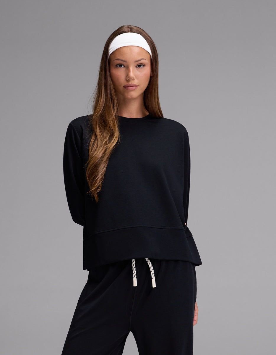 A woman with long brown hair and a white headband wears the Cozy Earth Women’s StudioLite Crewneck in Jet Black, paired with matching pants, standing against a plain gray background.|Color:Jet Black
