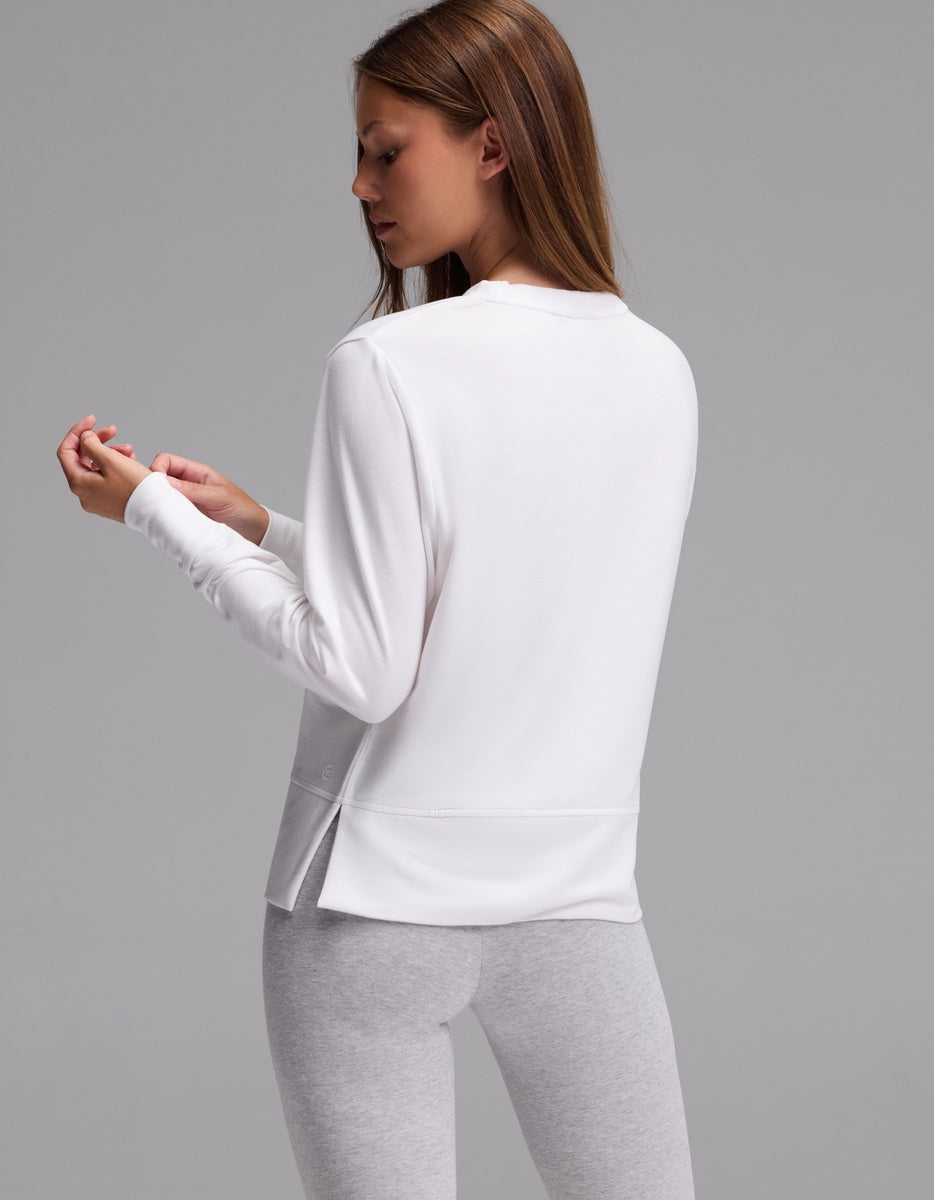 A woman with long brown hair is standing sideways against a plain gray background, wearing the Cozy Earth Women’s StudioLite Crewneck in white and light gray leggings, while looking at her hand.|Color:White