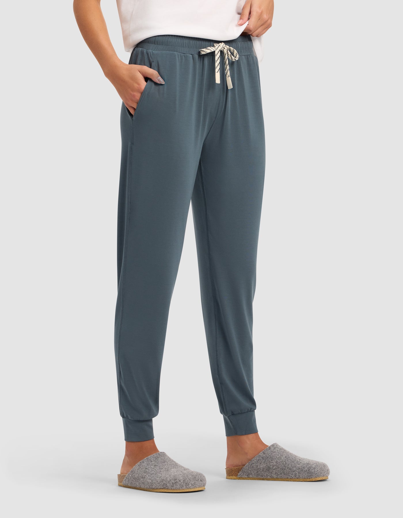 A person stands with hands in pockets, wearing Cozy Earth Women's Studio Jogger in teal with a white drawstring, a white top, and gray slip-on slippers against a plain light background.