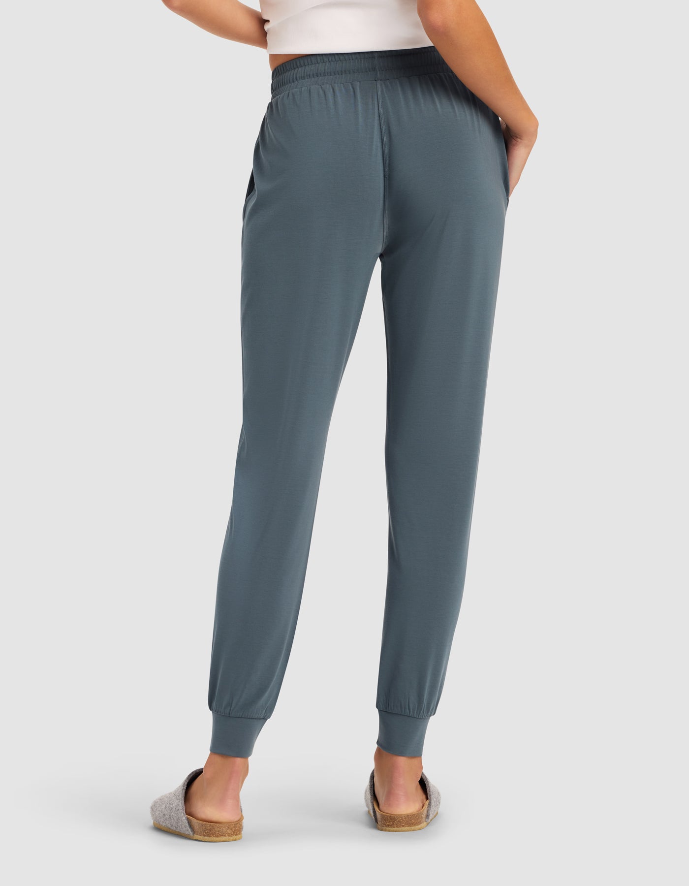 A person in Cozy Earth's Women's Studio Jogger, a white top, and gray slippers stands with their back to the camera, one hand on their hip.
