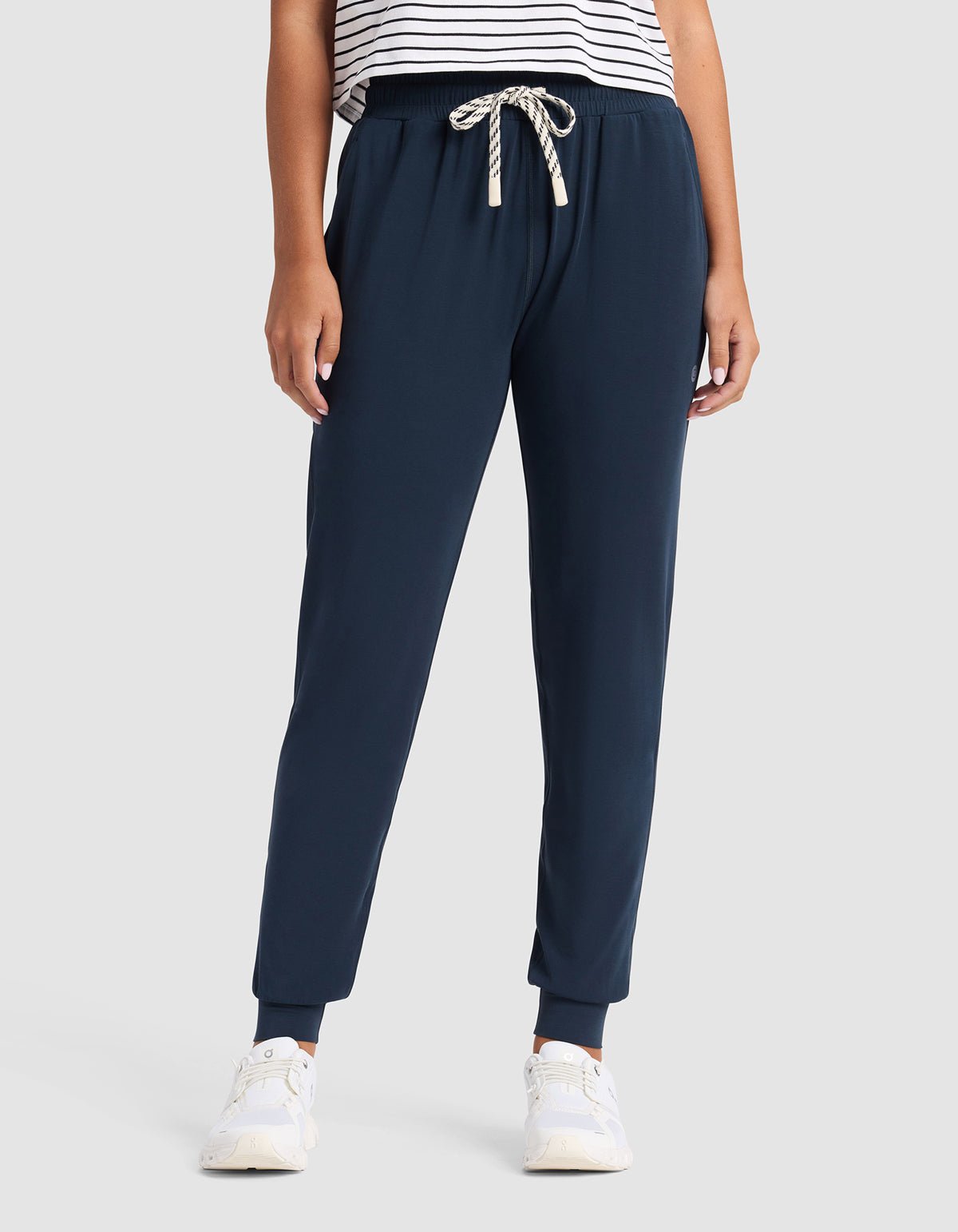 A person models Cozy Earth Women's Studio Jogger in navy blue with a white drawstring, paired with a white and black striped shirt and white sneakers, against a plain light gray background. The individual's head is not visible.