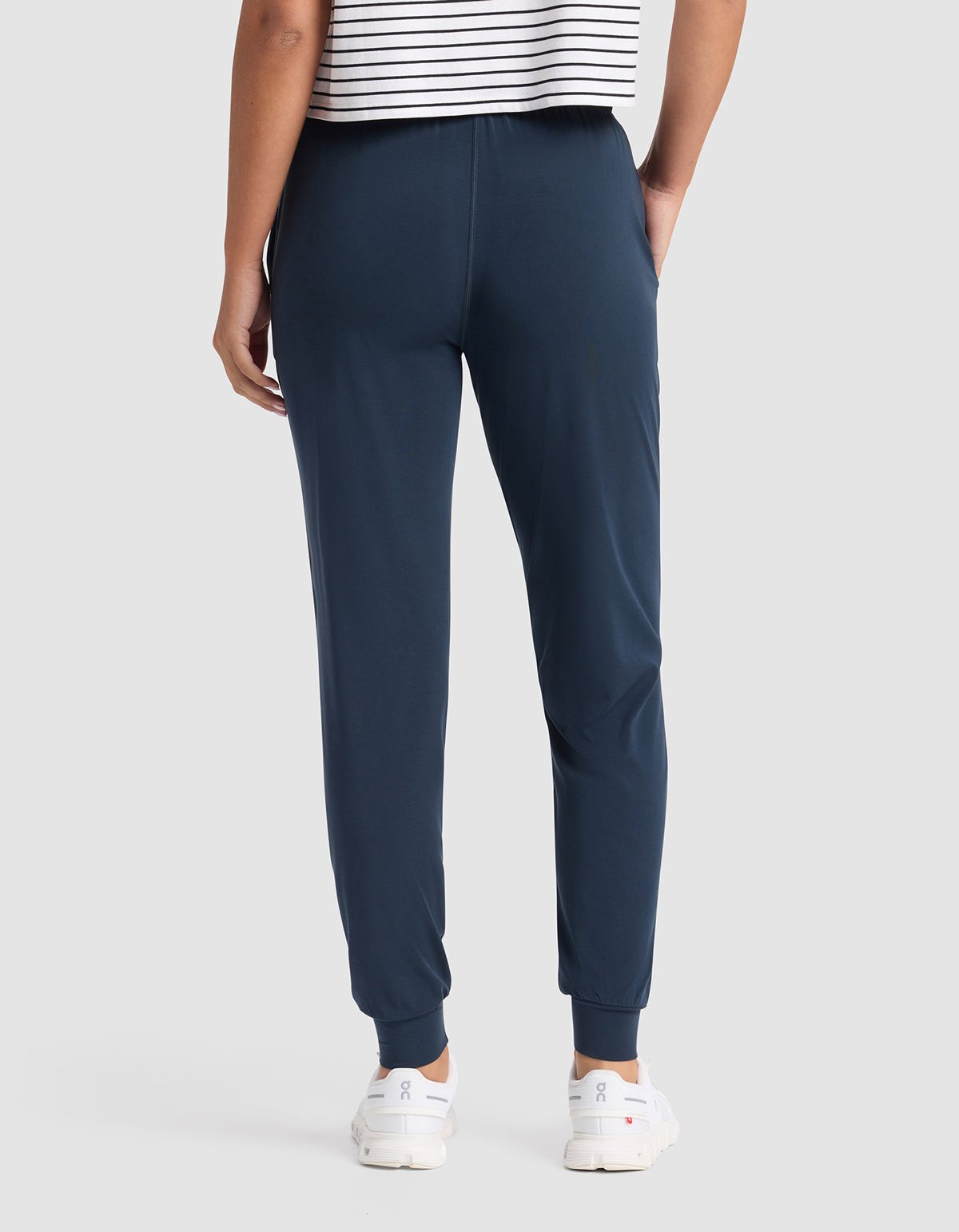 A person is shown from the back wearing Cozy Earth Women's Studio Jogger pants in navy, paired with a white and black striped shirt and white sneakers, against a plain light gray background.