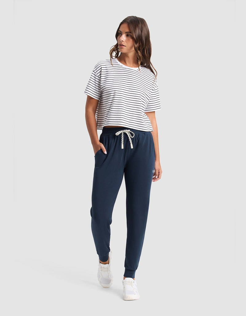 A woman with wavy brown hair stands against a plain background wearing Cozy Earth’s Women's Studio Jogger in navy blue, a white and black striped t-shirt, and white sneakers. Her left hand is in her pocket.