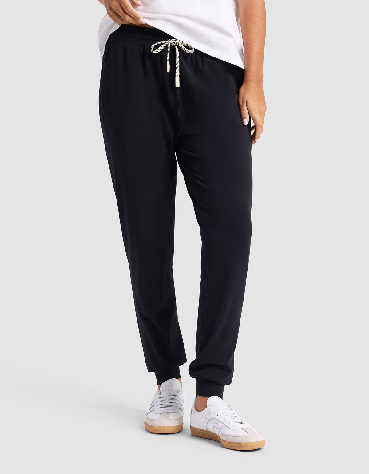 A person wearing Cozy Earth's Women's Studio Jogger in black with a white drawstring, a white T-shirt, and white sneakers stands against a plain light gray background.