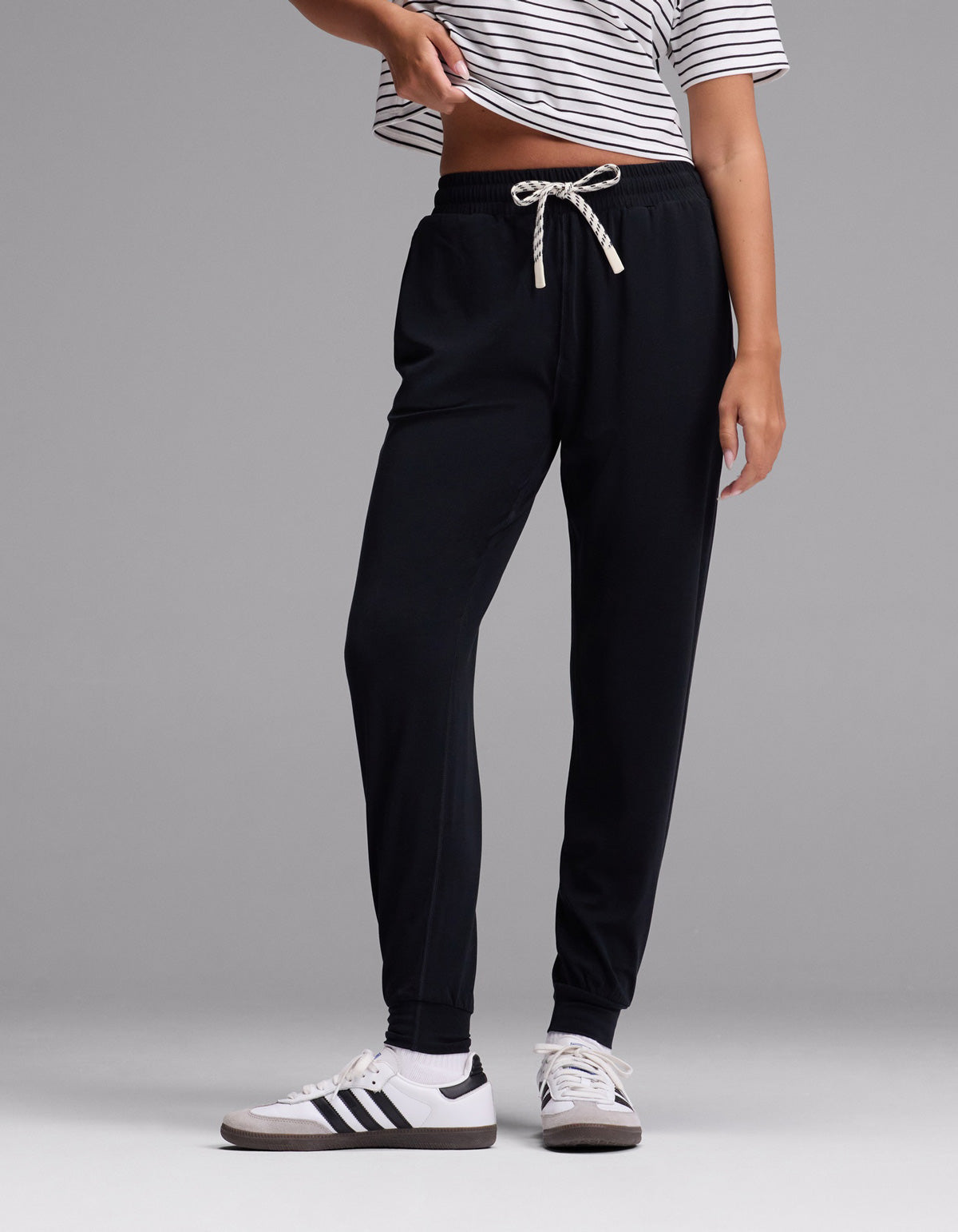 A person wearing a striped short-sleeve shirt, white sneakers with black stripes, and the Cozy Earth Women’s Studio Jogger in Jet Black stands against a plain gray background.|Color:Jet Black