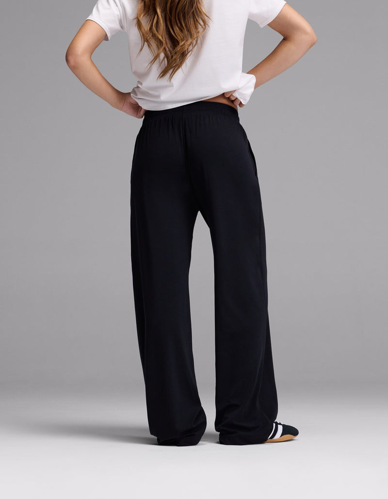 A person stands with hands on hips, facing away, wearing a white short-sleeve shirt and Cozy Earth Women’s Studio Wide Leg Pant in black. The background is plain gray.