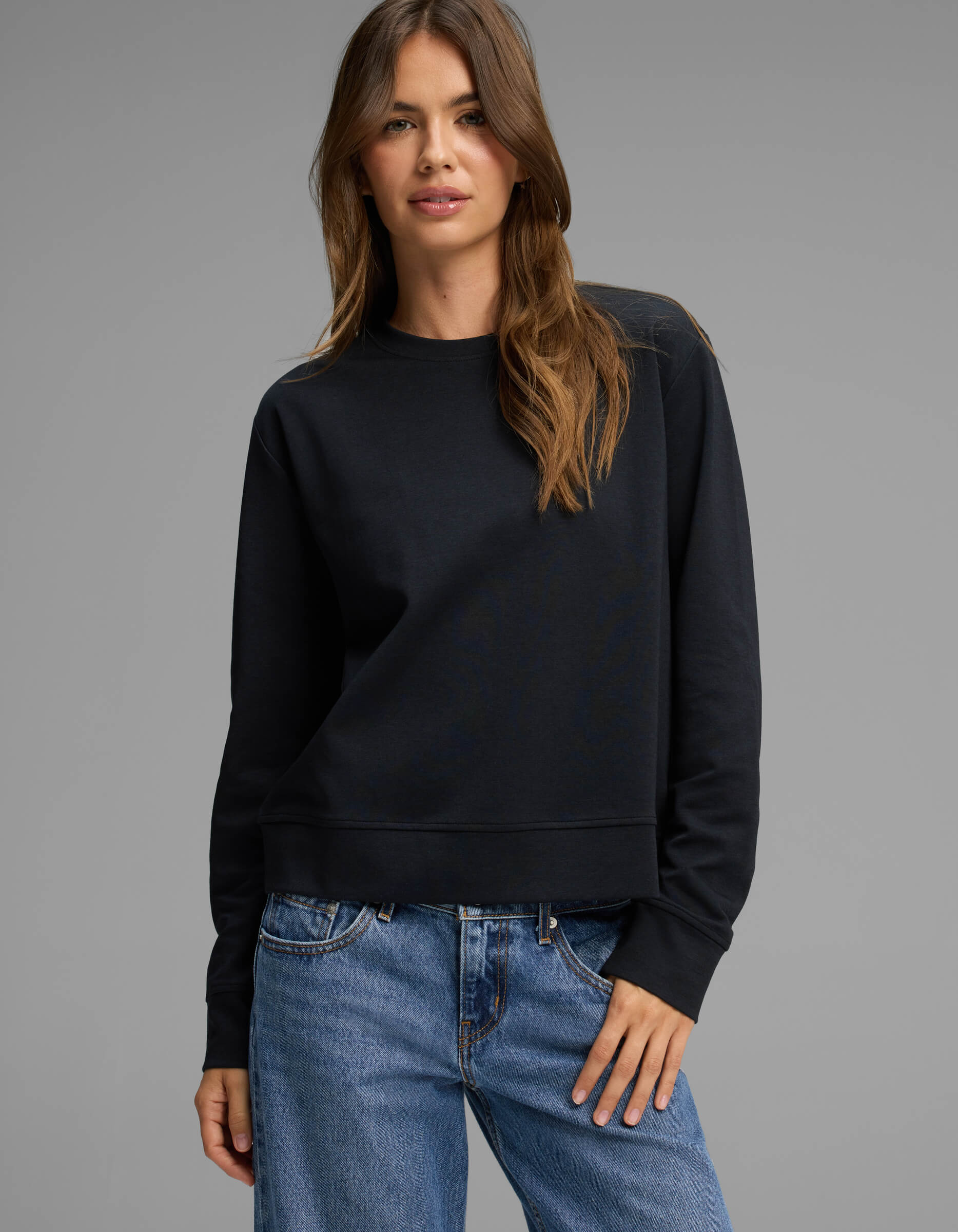 A woman with long brown hair stands against a plain gray background, looking at the camera with a slight smile while wearing Cozy Earth's Women's Versa Crewneck in black and blue jeans.|Color:Jet Black