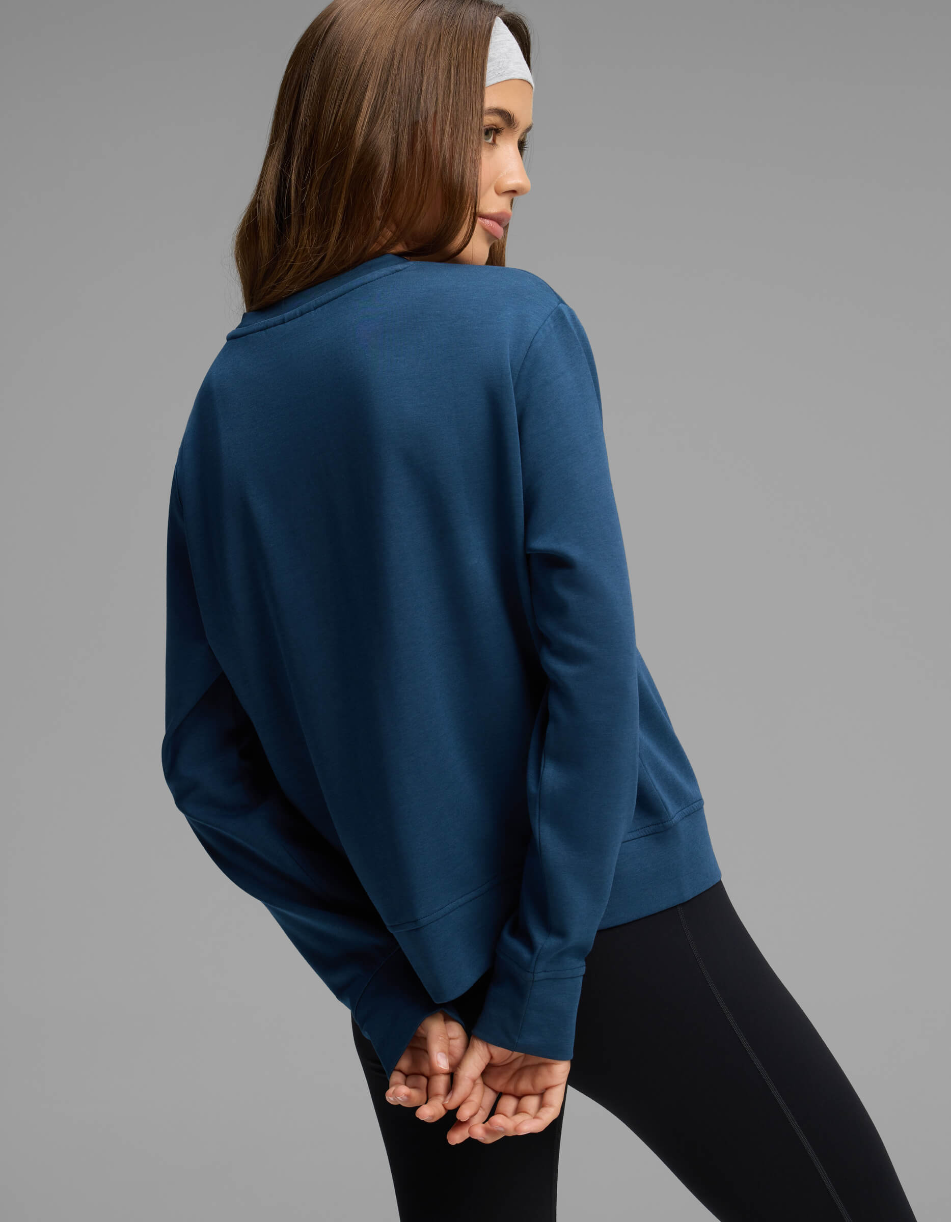 A woman stands with her back turned and hands clasped behind her against a plain gray background, wearing the Cozy Earth Women's Versa Crewneck in blue.