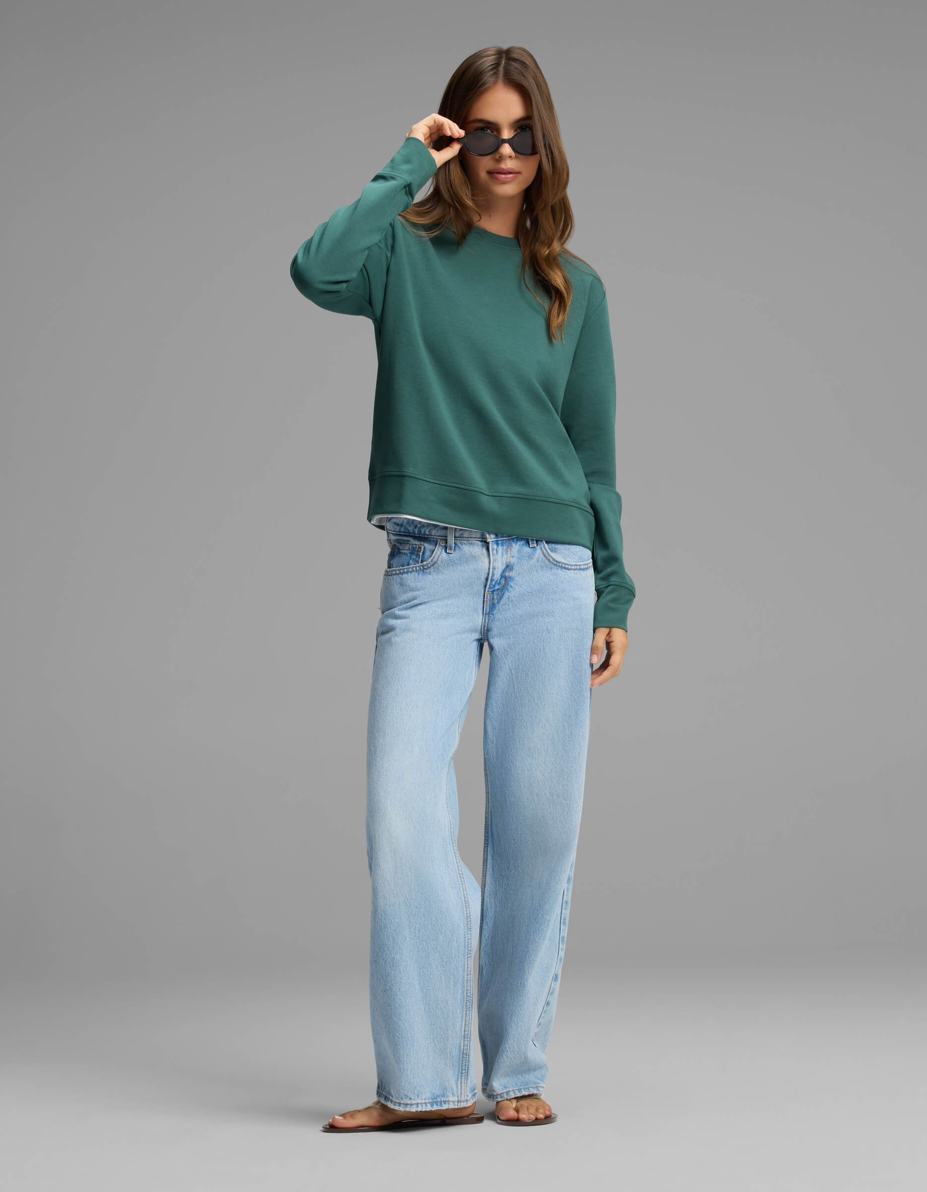 A woman stands against a plain gray background, looking at the camera while adjusting her sunglasses. She is wearing the Cozy Earth Women's Versa Crewneck in green, paired with light blue wide-leg jeans and sandals.