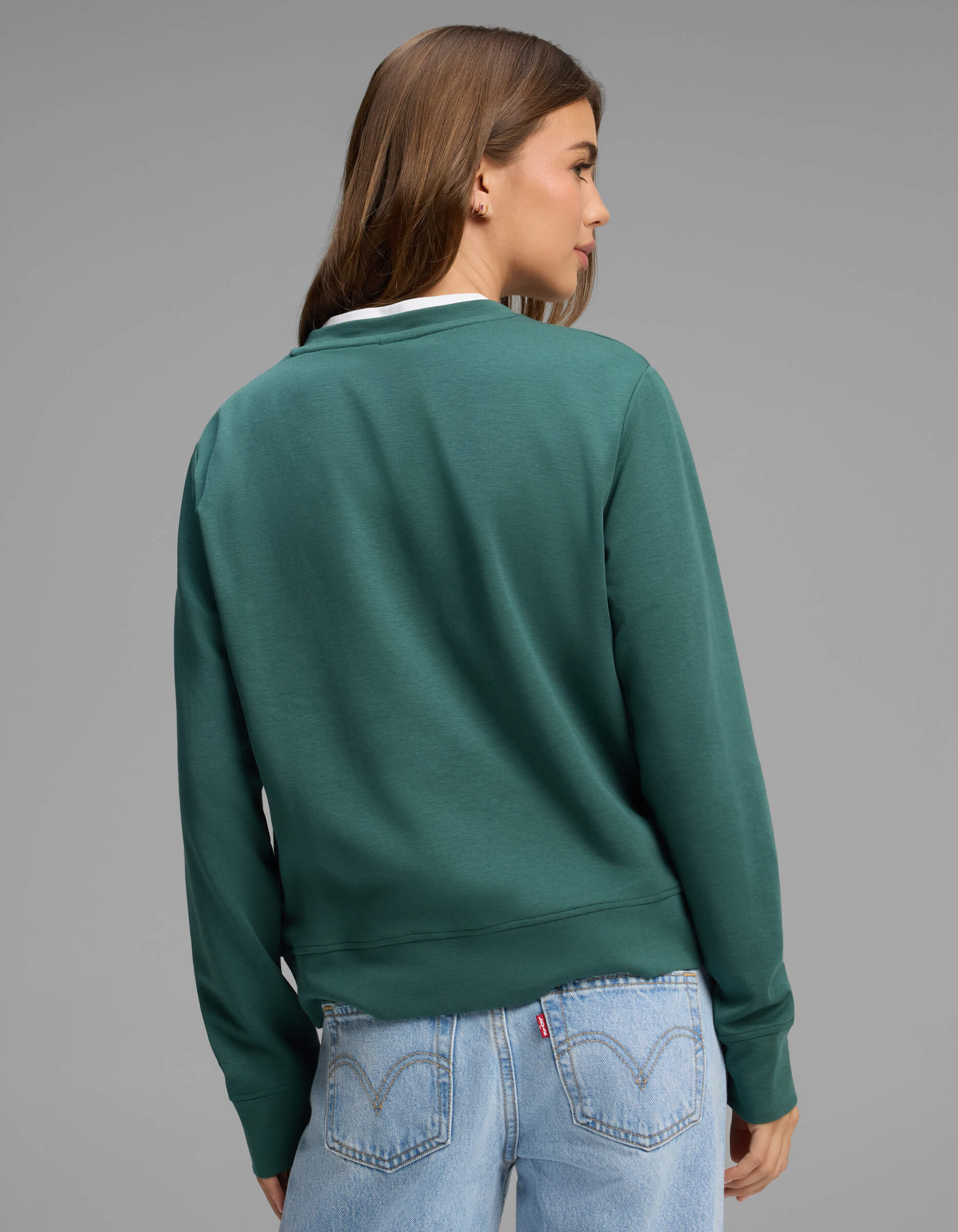 A woman with long brown hair, dressed in the Cozy Earth Women's Versa Crewneck in green and light blue jeans, stands facing away from the camera against a plain gray background.
