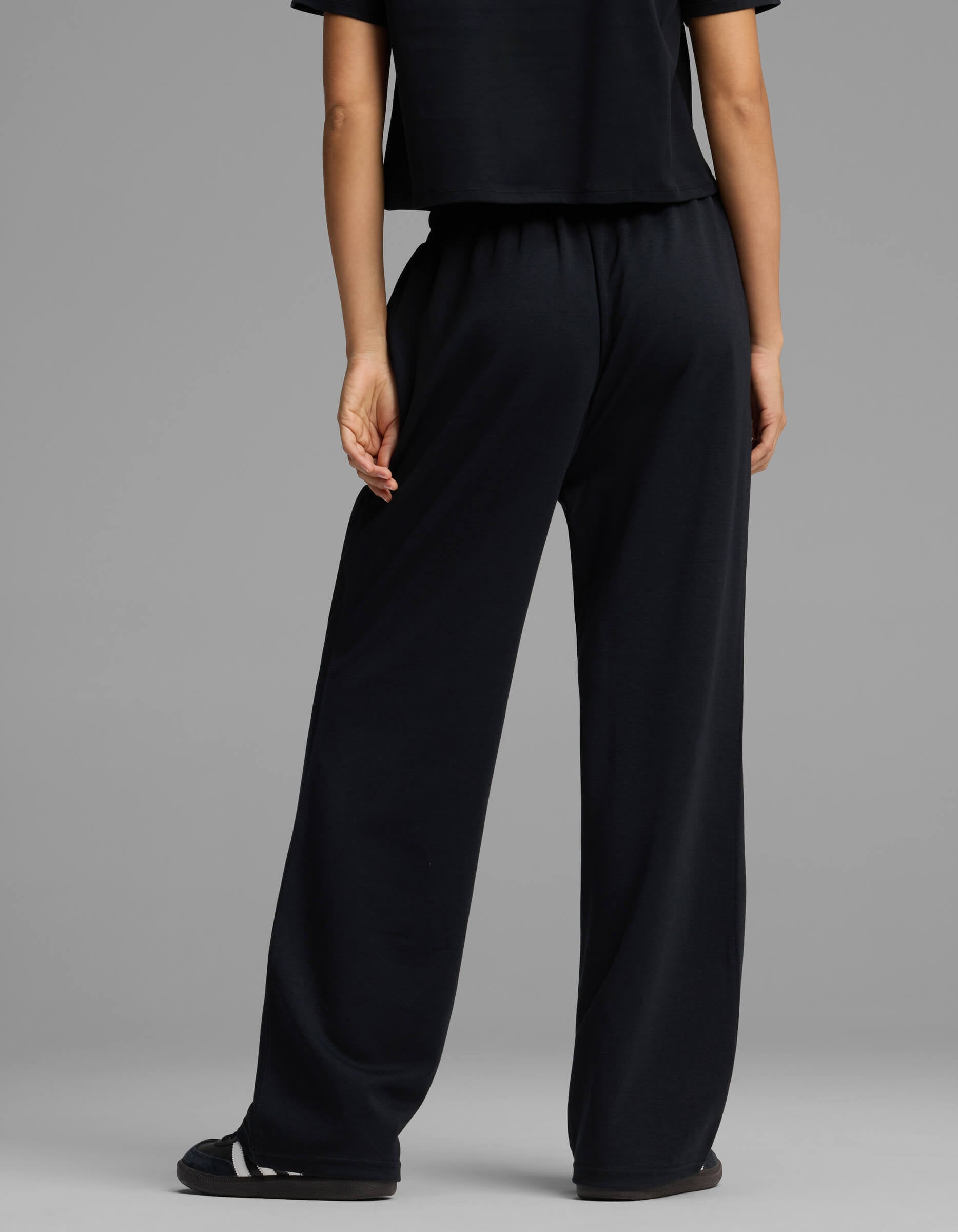 A person stands facing away, wearing Cozy Earth's Women's Versa Straight Leg Pant in black. The background is plain gray, and only the lower half of their body is visible.