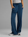 A person is wearing the Cozy Earth Women's Versa Straight Leg Pant in blue, paired with a gray tank top and gray sneakers, standing with hands in their pockets against a plain gray background.|Color:Obsidian