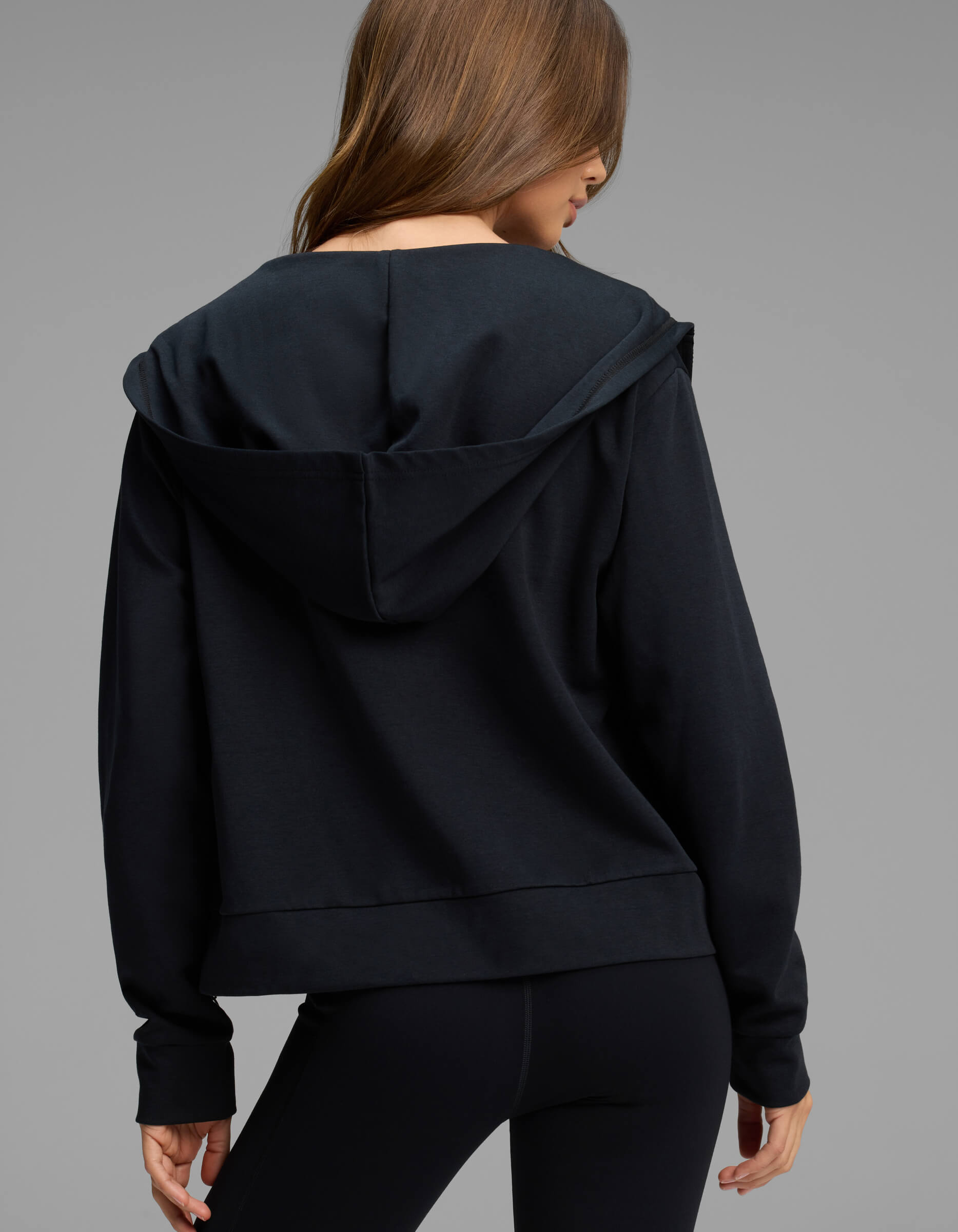 A person with long brown hair wears the Cozy Earth Women's Versa Full-Zip Hoodie in black, paired with black leggings, and stands facing away from the camera against a plain gray background.
