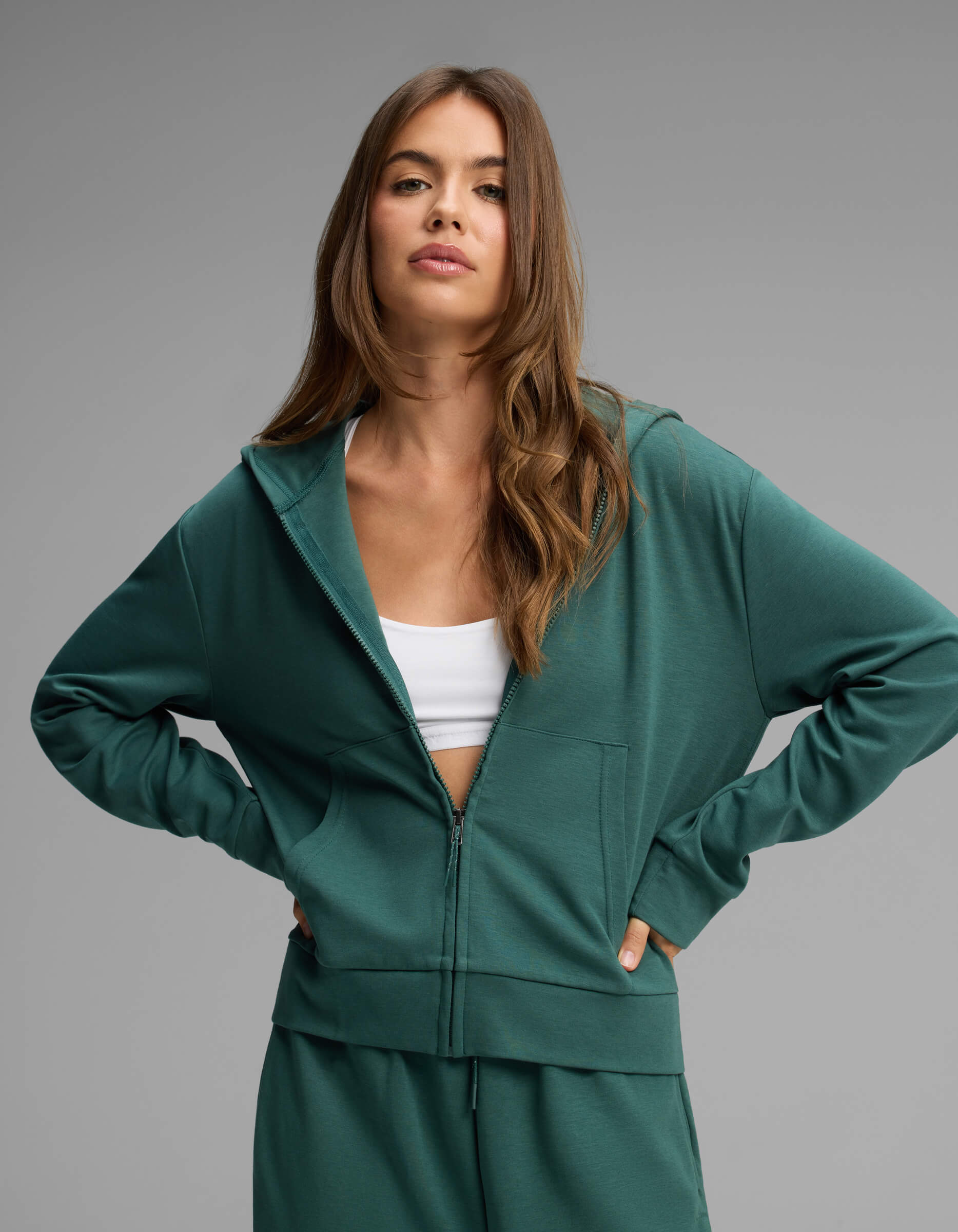 A woman with long brown hair stands with her hands on her hips against a plain gray background, wearing the Cozy Earth Women's Versa Full-Zip Hoodie in green over a white crop top.|Color:Uniform