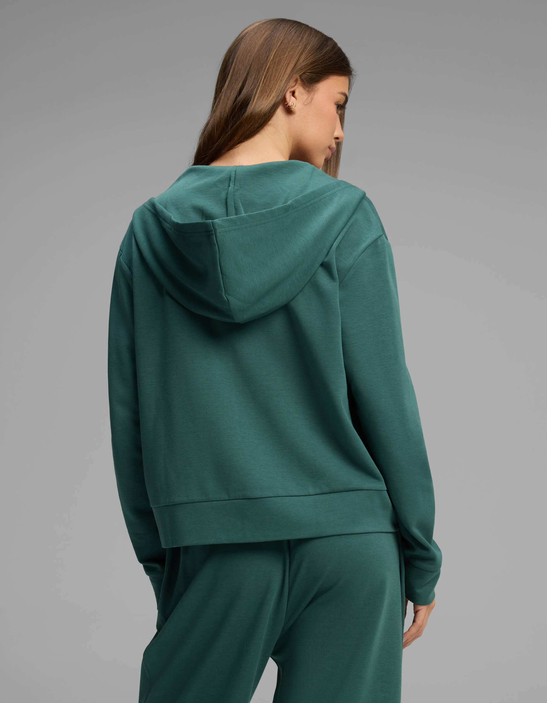 A woman with long brown hair is seen from behind against a plain gray background, wearing the Cozy Earth Women's Versa Full-Zip Hoodie in green with matching pants.|Color:Uniform