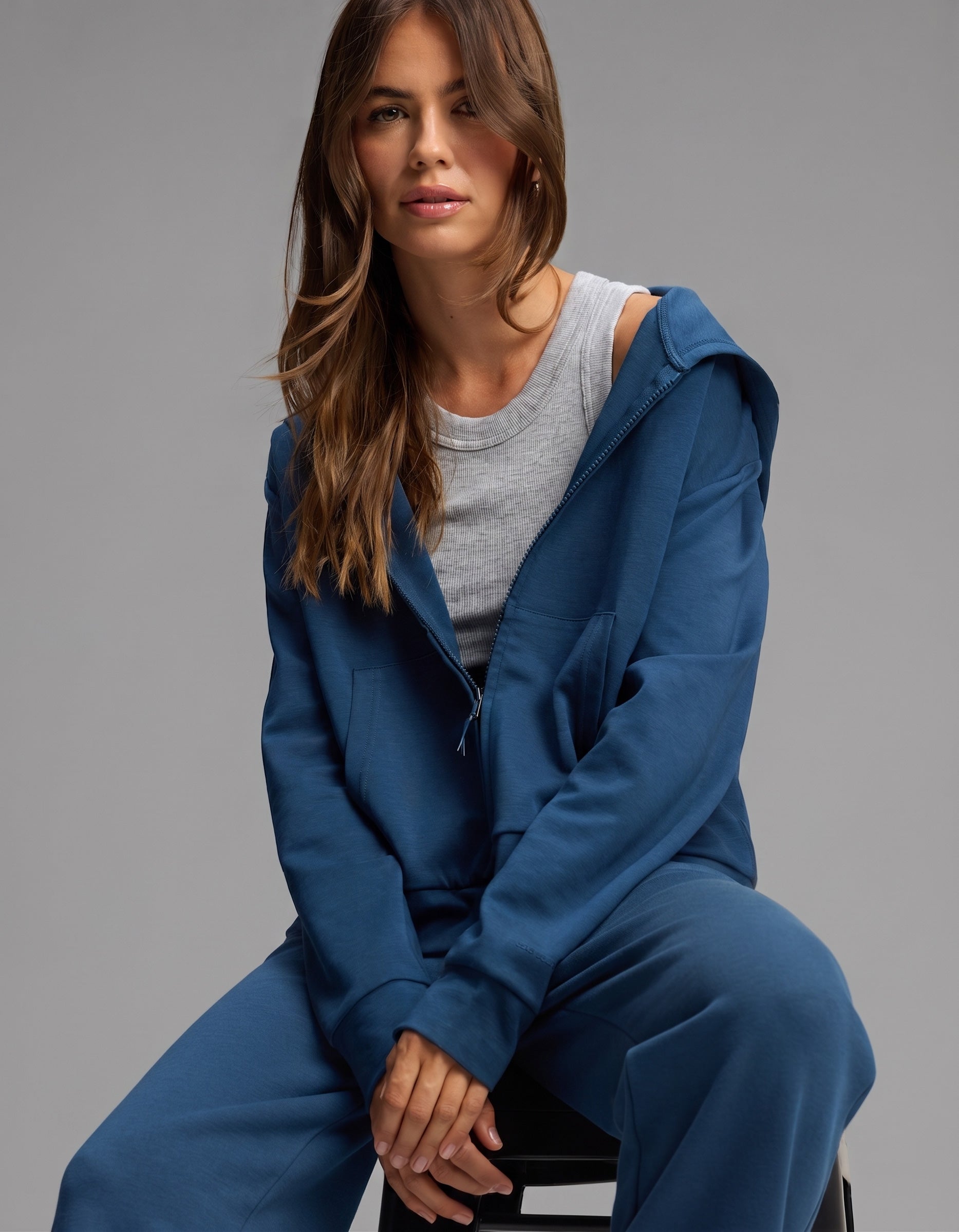 A woman with long brown hair sits on a stool against a plain gray background, wearing the Cozy Earth Women's Versa Zip-Down Hoodie & Straight Leg Pant Set in blue over a gray t-shirt.