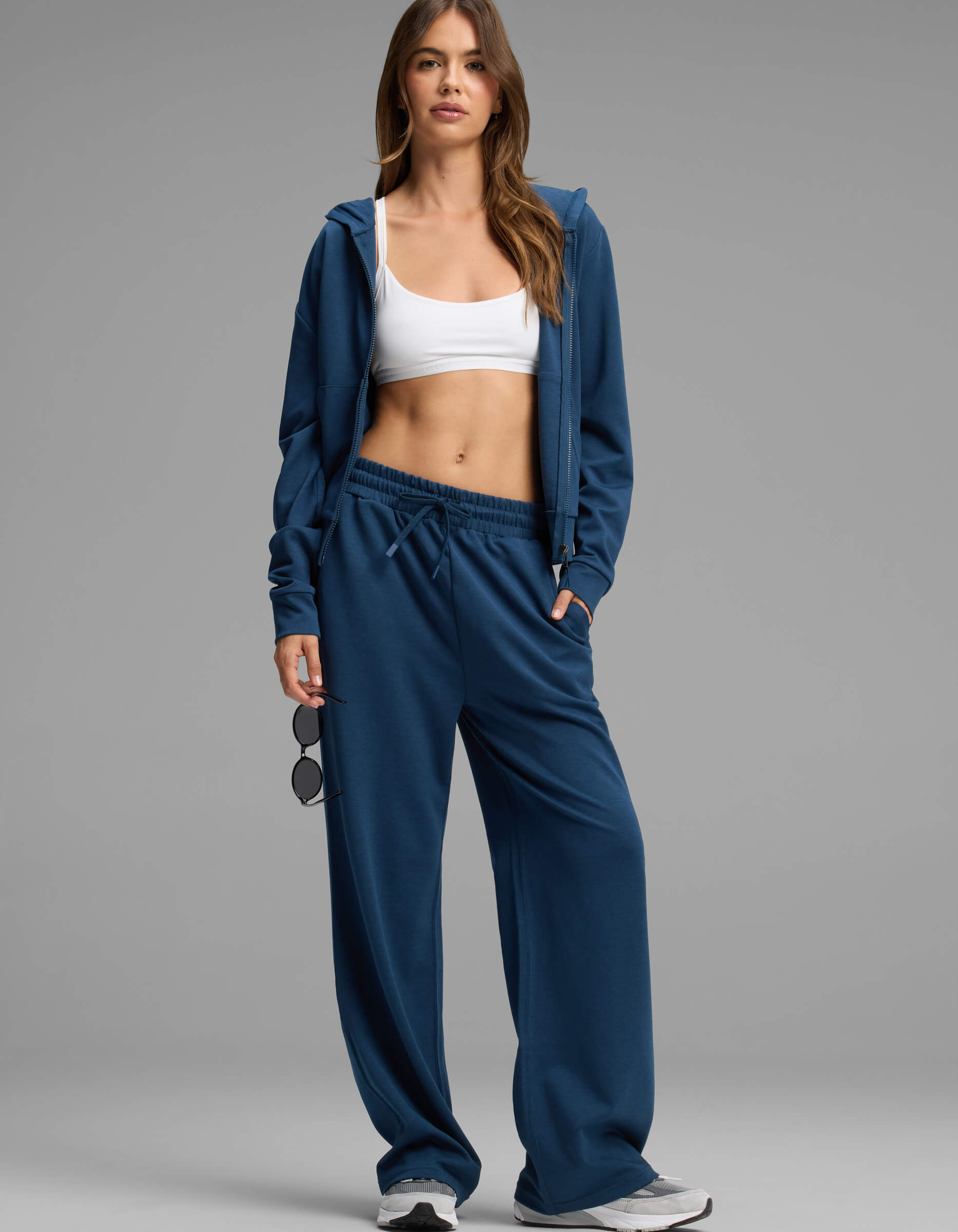 A woman stands wearing the Cozy Earth Women's Versa Zip-Down Hoodie & Straight Leg Pant Set in blue, paired with a white sports bra and sneakers. She holds sunglasses in one hand and gazes at the camera with a neutral expression against a plain background.|Color:Obsidian