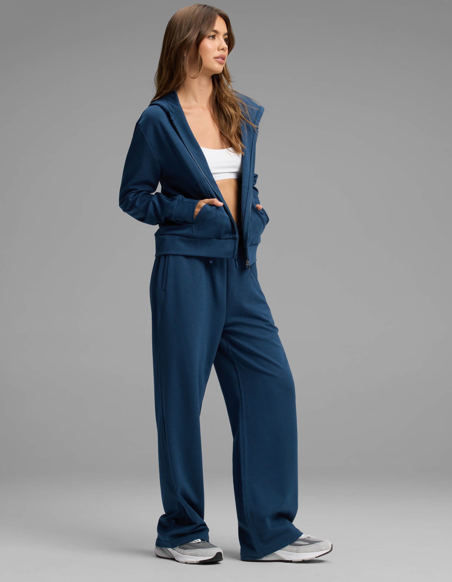 A woman stands against a plain gray background wearing the Cozy Earth Women's Versa Zip-Down Hoodie & Straight Leg Pant Set in blue, paired with a white crop top and white sneakers.