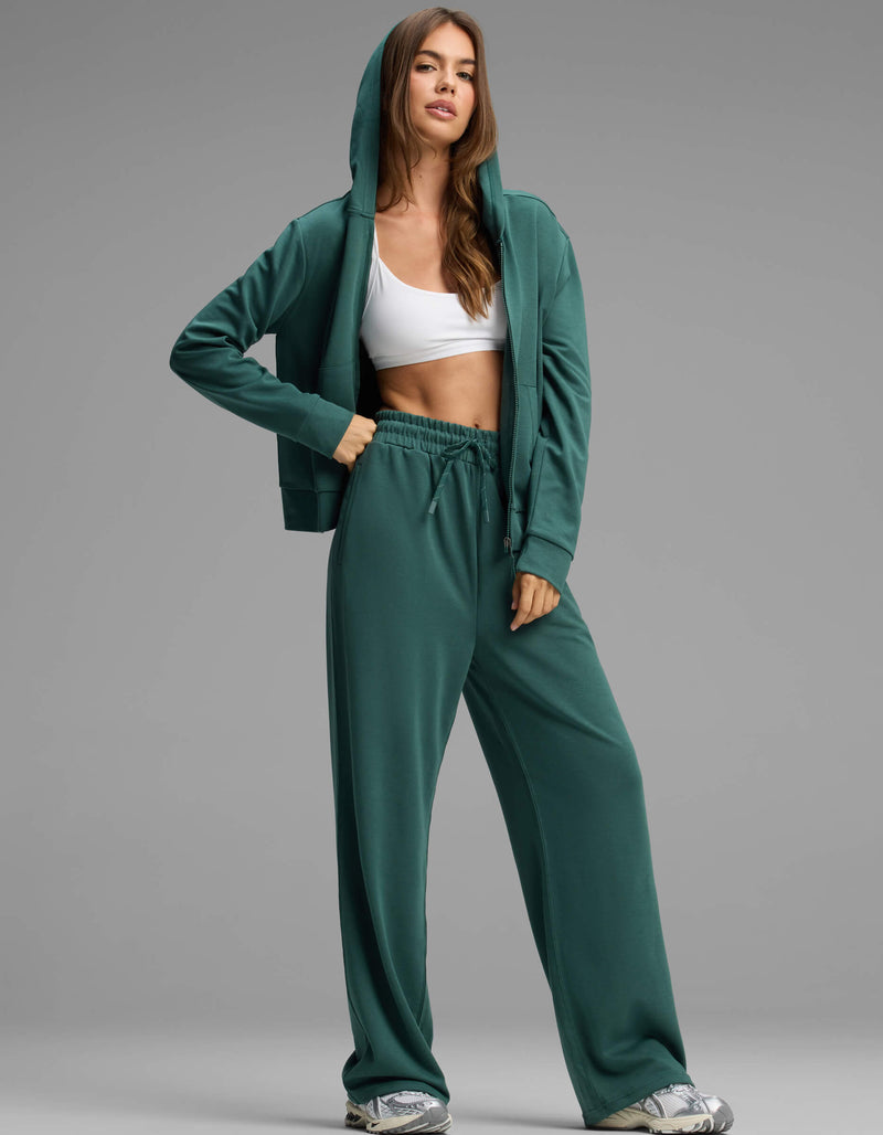 A woman stands confidently against a plain gray background, wearing Cozy Earth's Women's Versa Zip-Down Hoodie & Straight Leg Pant Set in green, paired with a white sports bra and sneakers.
