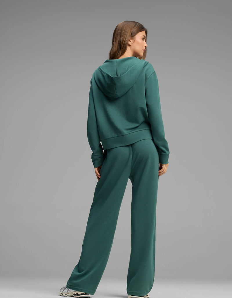 A person stands facing away, wearing the Cozy Earth Women's Versa Zip-Down Hoodie & Straight Leg Pant Set in green with white sneakers against a plain gray background.