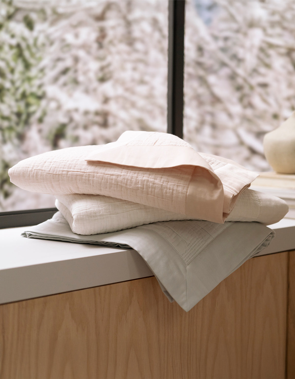 Three Cozy Earth Little Bee Matelasse Baby Blankets in soft pastels are neatly folded and stacked on a light wooden surface by a window, with blurred snow-covered trees visible outside.