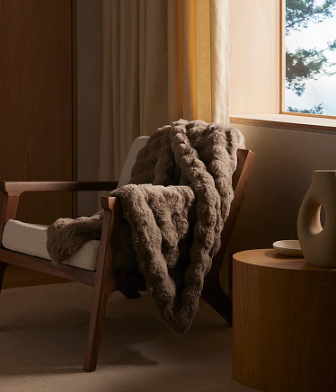 A wooden armchair draped with a textured gray throw, positioned beside a small round table near a sunlit window.