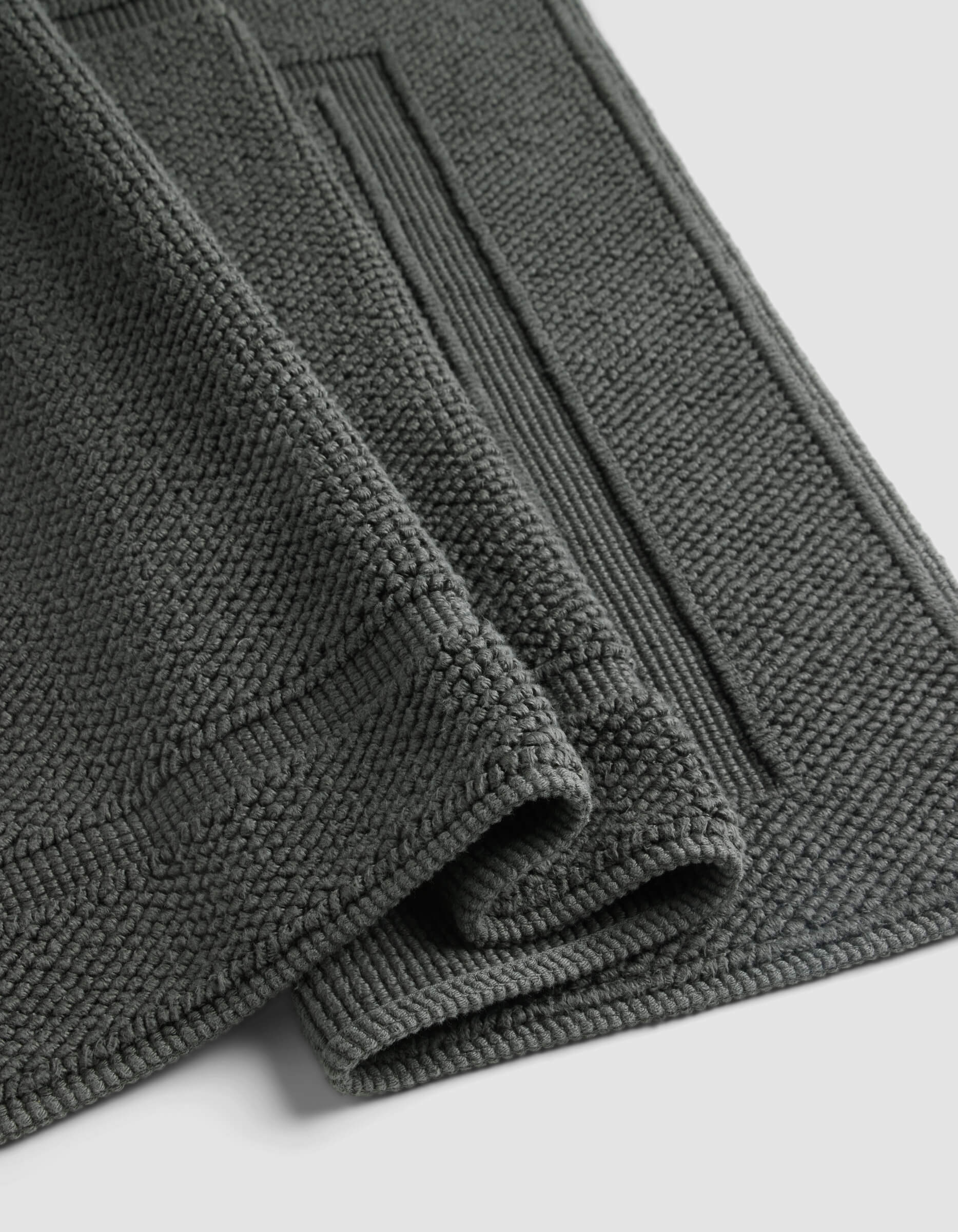The Cozy Earth Classic Bath Mat, shown folded against a light background, features a dark grey textured finish with a thick, soft pile and a rectangular border pattern.