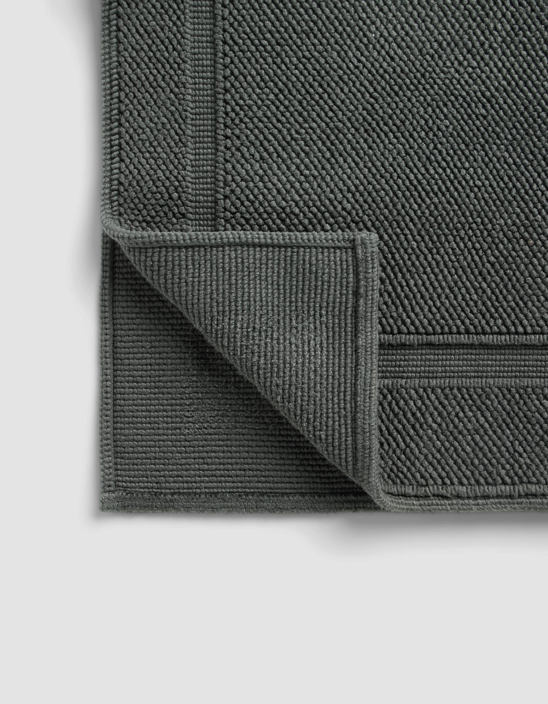 The Cozy Earth Classic Bath Mat, in dark gray with a textured woven pattern and bordered edges, is displayed on a light background with one corner folded to reveal the underside.
