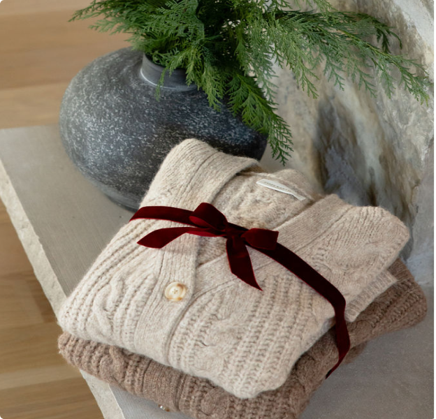 Two Cozy Earth Cable Knit Cardigans are folded and tied with a burgundy ribbon. They sit on a shelf near a vase holding evergreen boughs.