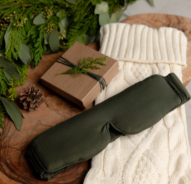 The Cozy Earth Adjustable Bamboo Sleep Mask is arranged near a small gift box, a cable knit stocking, and an evergreen bough.